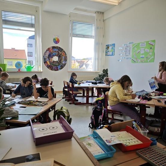 Ein Klassenzimmer mit mehreren Schülern, die an ihren Tischen arbeiten. Der Raum hat große Fenster und Poster an den Wänden. Eine Frau steht in der Nähe des Fensters, möglicherweise um die Schüler zu beobachten.