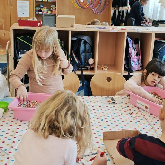 Eine Gruppe junger Kinder ist bei einer Bastelaktivität an einem Tisch mit Tupfentischdecke beschäftigt. Ein Kind schüttet Streusel aus einem rosa Behälter. Dahinter steht ein Holzlager mit verschiedenen Gegenständen.