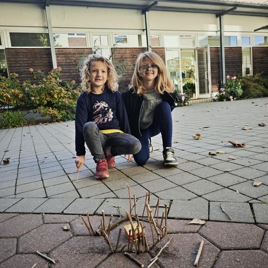 Zwei junge Mädchen hockten sich auf einem Ziegelweg vor einem Gebäude, lächelten und hielten Stöcke. Vor ihnen befand sich ein kleiner, selbstgebauter Feuerplatz aus Zweigen.