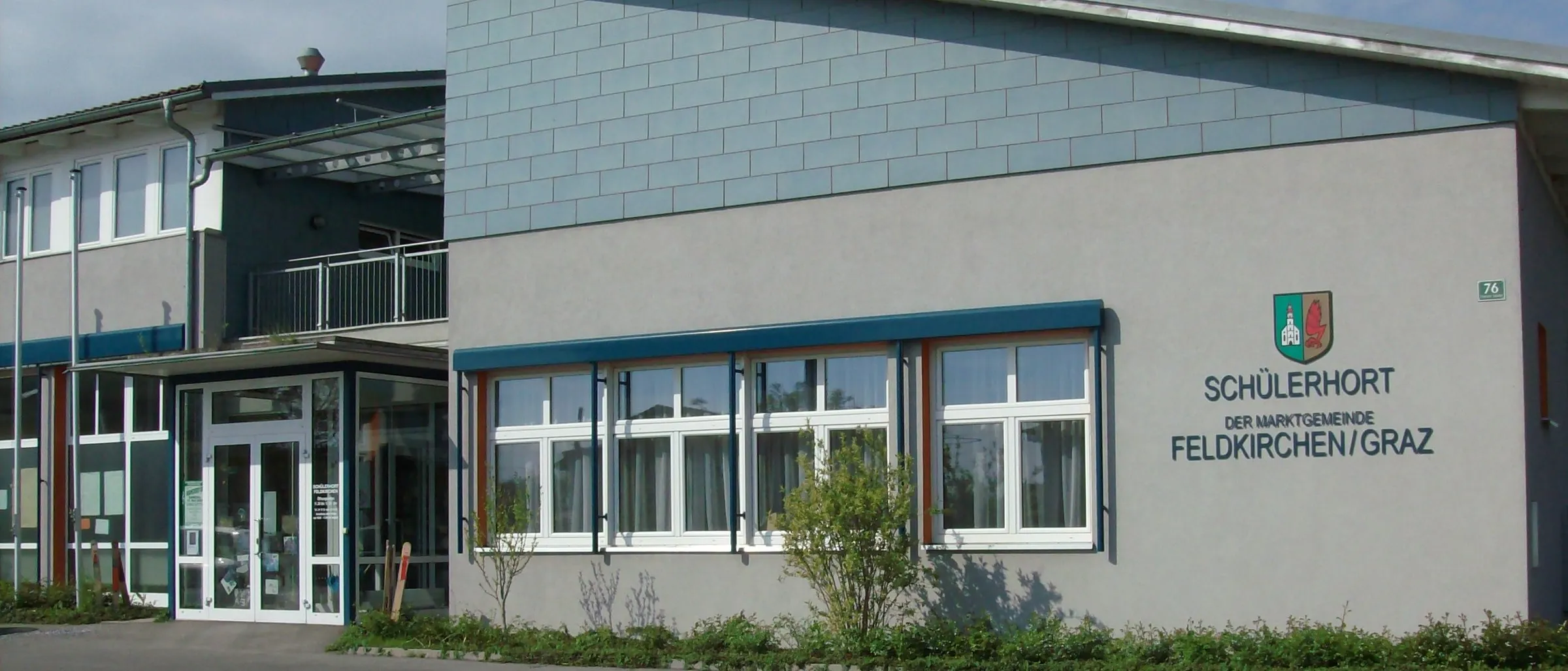 Der Schulerhof der Gemeinde Feldkirchen/O hat eine blaue Wand mit Fenstern und einem Logo. Das Gebäude verfügt über einen Balkon und einen Haupteingang.