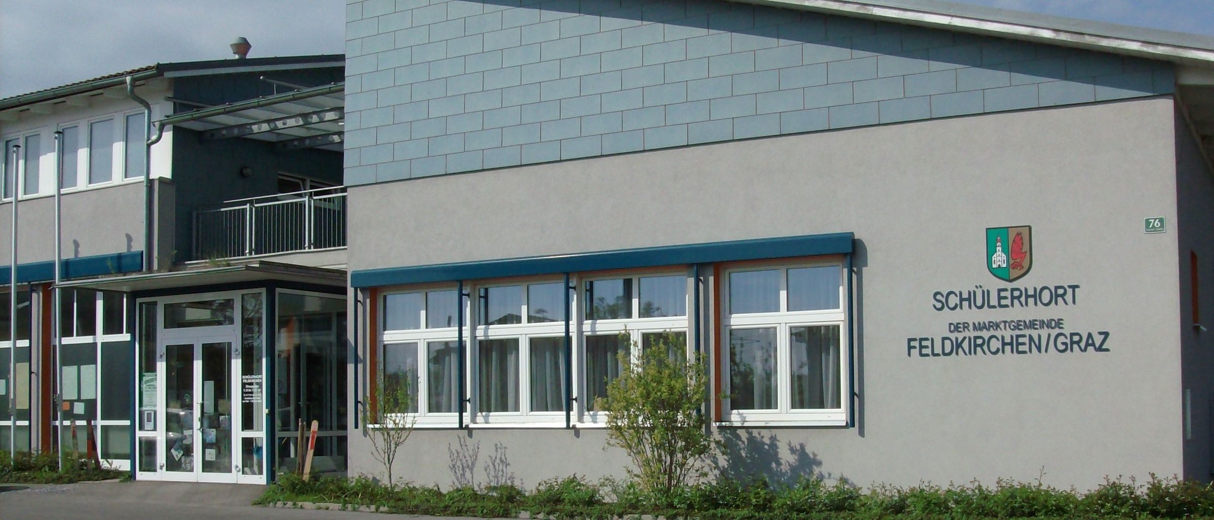 Der Schulerhof der Gemeinde Feldkirchen/O hat eine blaue Wand mit Fenstern und einem Logo. Das Gebäude verfügt über einen Balkon und einen Haupteingang.