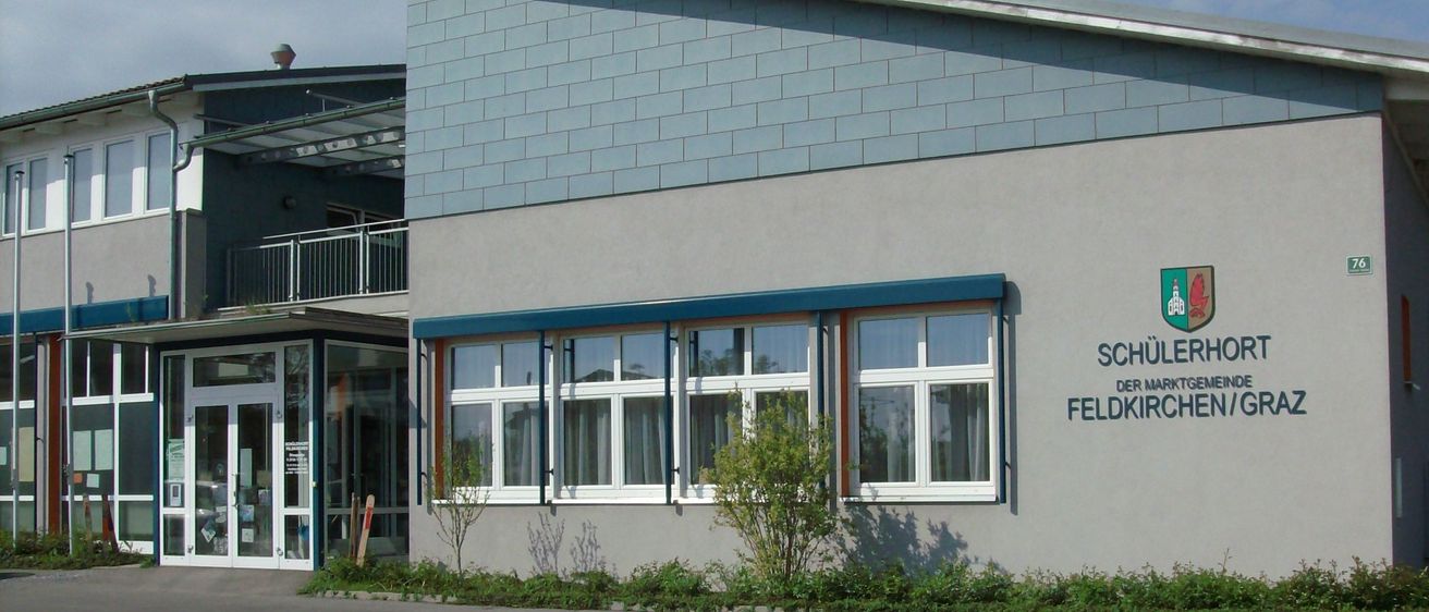 Der Schulerhof der Gemeinde Feldkirchen/O hat eine blaue Wand mit Fenstern und einem Logo. Das Gebäude verfügt über einen Balkon und einen Haupteingang.