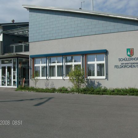 Vorderansicht eines Gebäudes mit blauem Dach, weißen Fenstern und einem Schild, das 'Schiilerhorp der Marktgemeinde Feldkirchen/O' liest.
