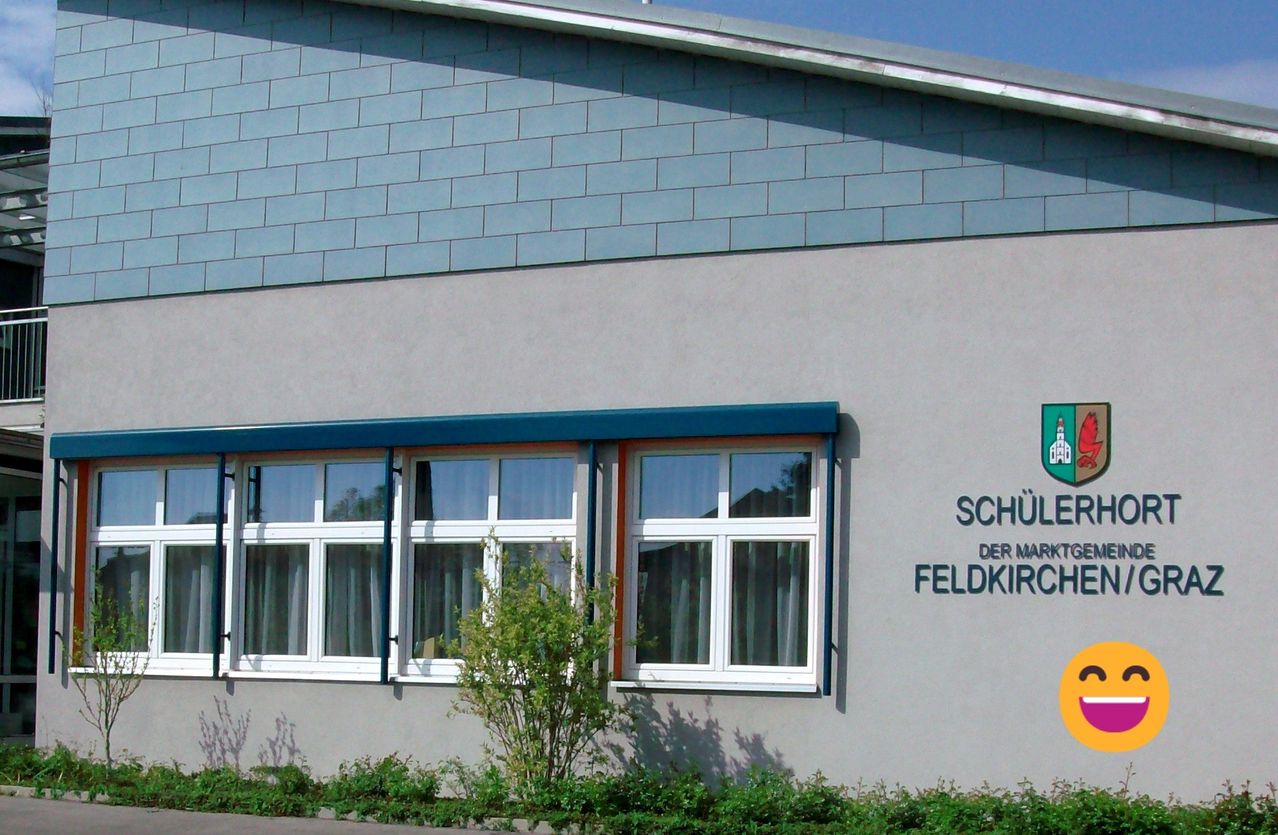 Die Vorderseite eines Schulgebäudes mit grünem Rand und weißen Fenstern, ein grünes Schild mit der Aufschrift 'Schul der Mark Feldkirch'.