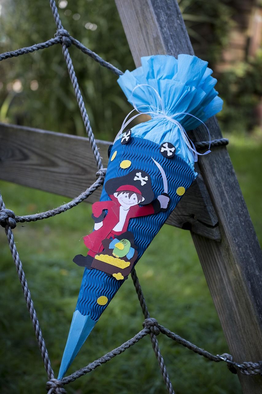 Ein blauer kegelförmiger Geschenkbeutel mit Piraten-Thema, der einen gezeichneten Piratencharakter zeigt, hängt an einer Holzstruktur mit Seilen. Der Beutel ist mit Totenkopf und Kreuzknochen und Goldmünzen verziert.