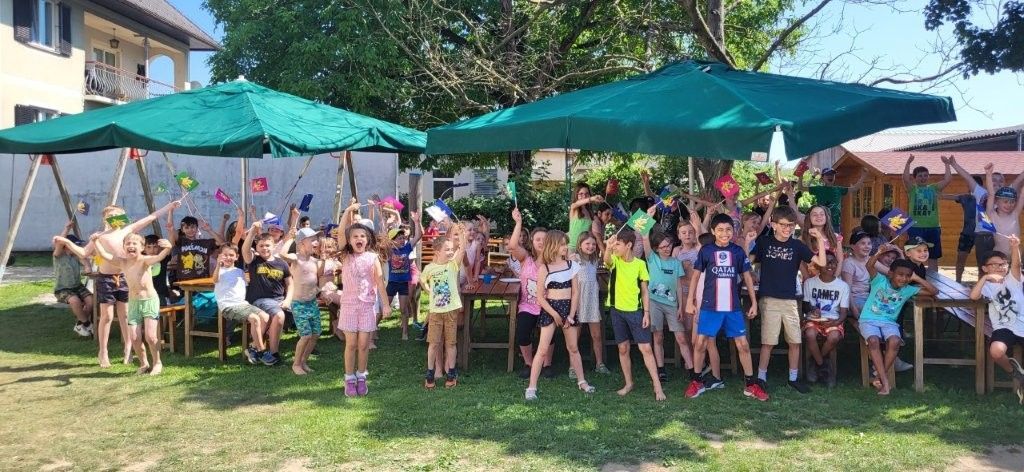 Eine Gruppe von Kindern und Erwachsenen versammelt sich unter grünen Schirmen in einem Rasenbereich. Sie halten Flaggen und scheinen für ein Foto zu posieren.