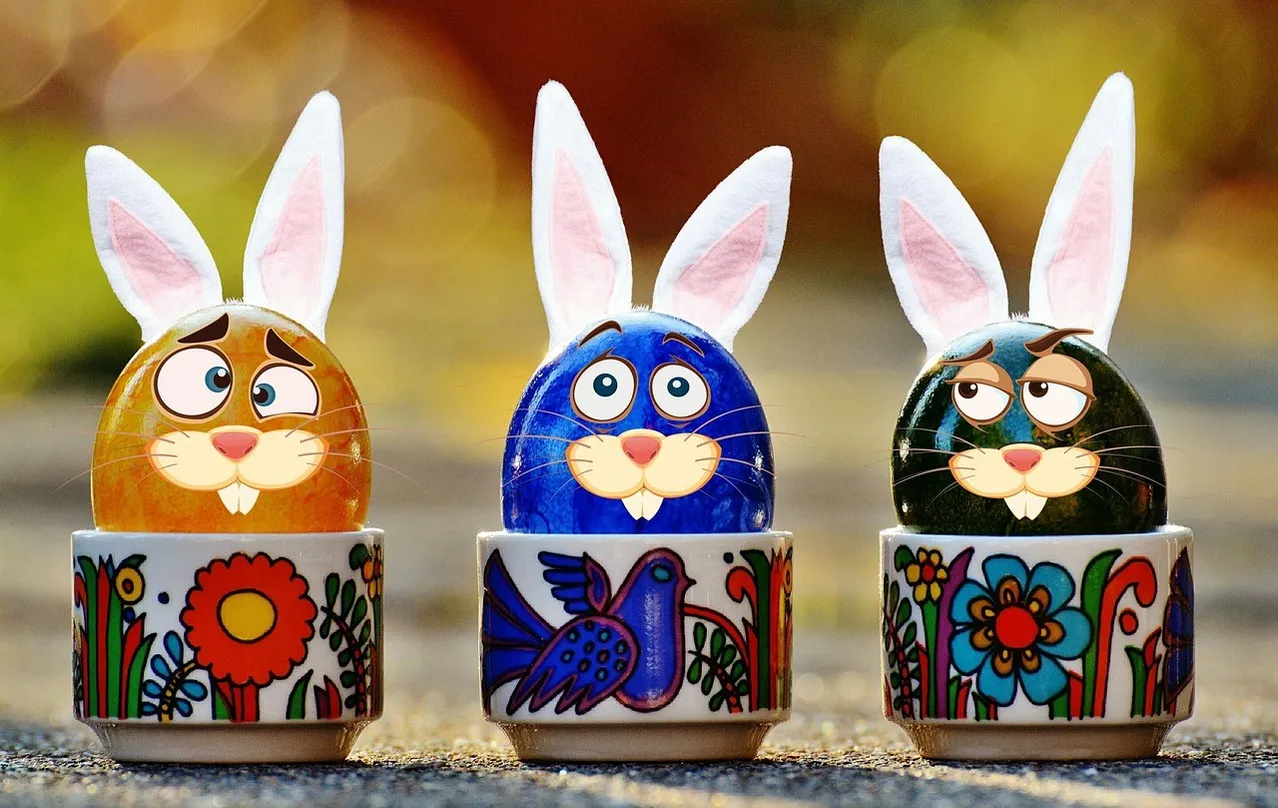 Drei bunte Keramikeier mit Hasenohren. Jedes Ei ist in einer anderen Farbe bemalt und hat ein anderes Design auf seinem Becher. Die Eier liegen auf einer Betonoberfläche.
