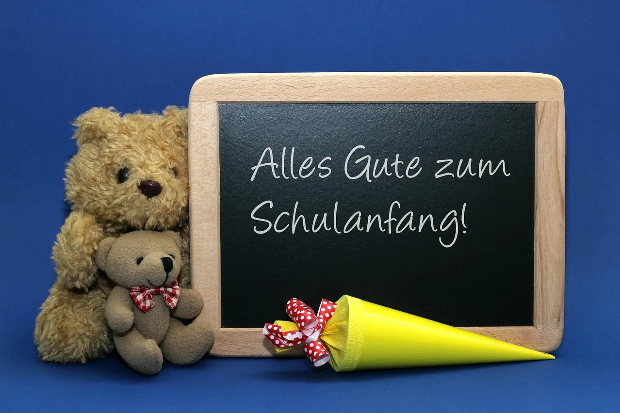 Eine Tafel mit der Aufschrift 'Alles Gute zum Schulanfang!' ist vor einem blauen Hintergrund zu sehen. Zwei Teddybären, einer mit einer Schleife, stehen neben der Tafel. Ein gelber Kegel mit einem roten und weißen Band ist ebenfalls vorhanden.