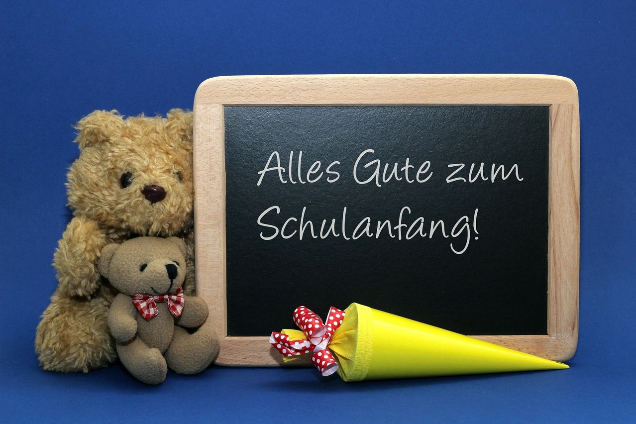 Eine Tafel mit der Aufschrift 'Alles Gute zum Schulanfang!' ist vor einem blauen Hintergrund zu sehen. Zwei Teddybären, einer mit einer Schleife, stehen neben der Tafel. Ein gelber Kegel mit einem roten und weißen Band ist ebenfalls vorhanden.