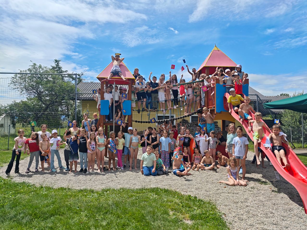 Eine große Gruppe von Kindern versammelt sich auf einem Spielplatz. Einige stehen auf den Rutschen, andere sitzen auf dem Boden. Sie alle lächeln für ein Foto.