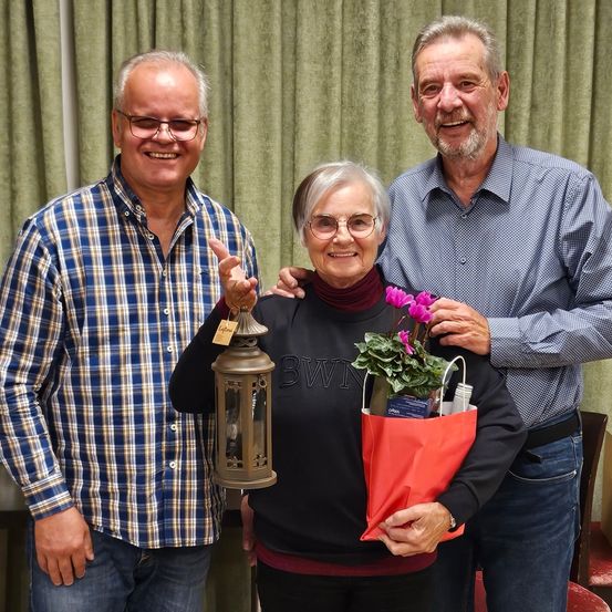 Drei Menschen stehen lächelnd in einem Raum. Eine Frau in der Mitte hält eine Laterne und einen Blumenstrauß. Der Mann rechts von ihr hat seine Hand auf ihrer Schulter. Hinter ihnen ist ein grüner Vorhang.