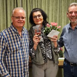 Drei Menschen stehen eng beieinander und lächeln. Die Frau hält Blumen und Schokolade, während die Männer Brillen und Gürtel tragen.