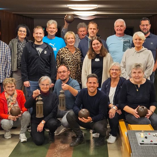 Eine Gruppe von Menschen versammelt sich zu einem Foto, einige halten Bowlingkugeln, in einer Bowlingbahn. Sie tragen verschiedene Kleidungsstücke und einige tragen Brillen. Ein Tisch mit einem Bedienfeld befindet sich vorne.