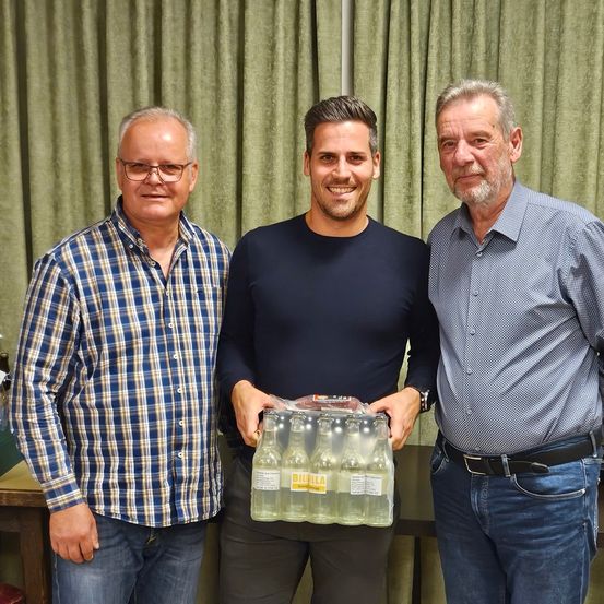 Drei Männer stehen zusammen, der in der Mitte hält einen Packen Flaschen und lächelt. Dahinter ist ein grüner Vorhang.