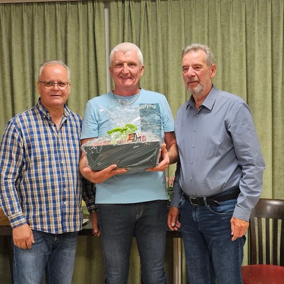 Drei Männer stehen nebeneinander vor einem grünen Vorhang. Der Mann in der Mitte hält eine Geschenkbox. Der Mann auf der linken Seite trägt ein blaues kariertes Hemd und der Mann auf der rechten Seite trägt ein blaues Polohemd.