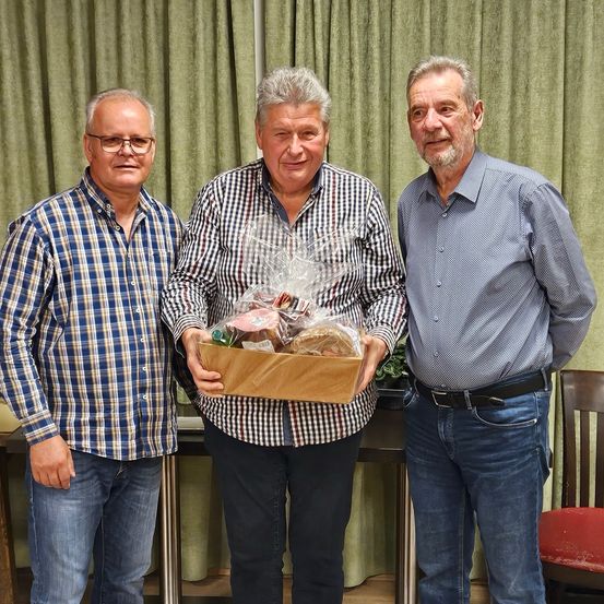 Drei Männer stehen zusammen; der in der Mitte hält eine Schachtel mit Essen. Sie lächeln und scheinen für ein Foto zu posieren. Hinter ihnen ist ein Vorhang und ein Stuhl zu sehen.