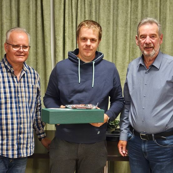 Drei Männer stehen zusammen und halten eine grüne Box, einer der Männer trägt eine blaue Kapuzenjacke und hält die Box, während die anderen beiden Männer neben ihm stehen und lächeln.