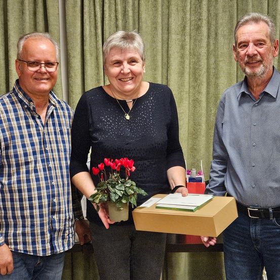Drei Menschen stehen lächelnd vor einem grünen Vorhang. Die Frau hält eine Topfpflanze und eine Schachtel. Der Mann links trägt ein kariertes Hemd, während der Mann rechts ein blaues Hemd trägt.