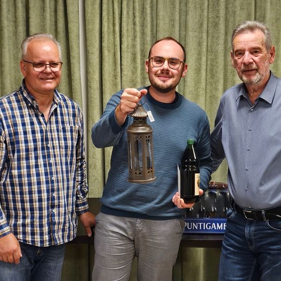 Drei Männer stehen gemeinsam vor einem grünen Vorhang. Der Mann in der Mitte hält eine Laterne und eine Flasche. Der Mann links trägt ein kariertes Hemd und der Mann rechts trägt ein blaues Hemd.