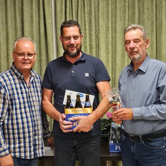 Drei Männer stehen nebeneinander vor einem grünen Vorhang. Der Mann in der Mitte hält eine Box Panther-Bier und einen Plastikbeutel. Der Mann auf der linken Seite trägt ein kariertes Hemd und der Mann auf der rechten Seite trägt ein blaues Langarmhemd.