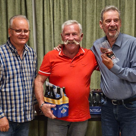 Drei Männer lächeln und posieren für ein Foto. Der Mann in der Mitte hält eine Schachtel Panther-Bier. Der Mann auf der rechten Seite hält eine Plastiktüte. Der Mann auf der linken Seite trägt ein kariertes Hemd.