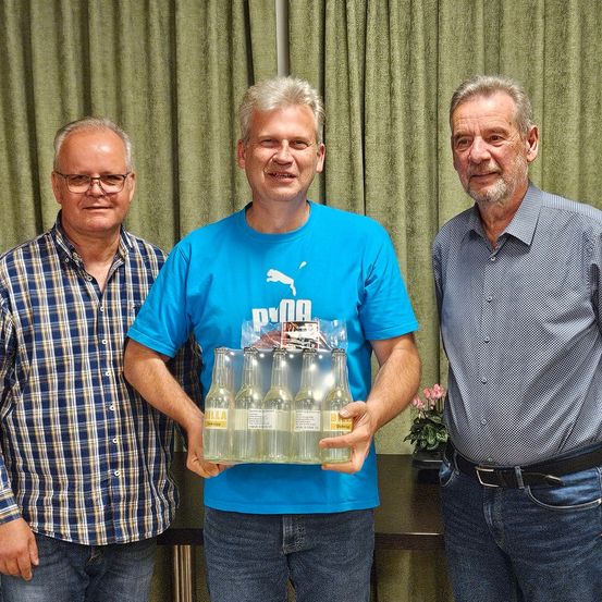 Drei Männer stehen lächelnd hinter einem Tisch. Der Mann in der Mitte hält eine Packung mit sechs klaren Flaschen. Der Mann auf der linken Seite trägt ein kariertes Hemd, und der Mann auf der rechten Seite trägt ein kariertes Hemd. Hinter ihnen befinden sich grüne Vorhänge und eine Pflanze.