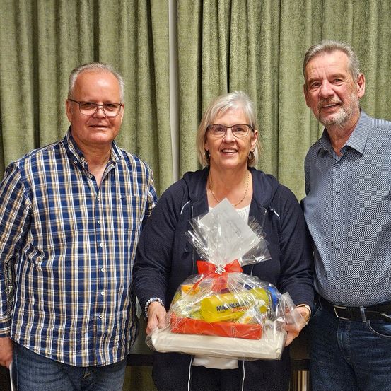 Drei Personen stehen lächelnd hinter einem Tisch. Eine Frau hält einen Geschenkkorb. Der Mann links trägt ein kariertes Hemd, der andere Mann ein blaues Hemd. Sie stehen vor grünen Vorhängen.