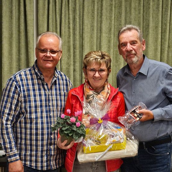Drei Menschen stehen eng beieinander und lächeln. Die Frau in der Mitte hält einen Geschenkkorb mit einer Pflanze und einer Karte. Der Mann auf der rechten Seite hält eine Plastiktüte. Der Mann auf der linken Seite trägt eine Brille und ein kariertes Hemd.
