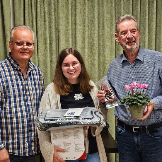 Drei Personen stehen zusammen, zwei Männer und eine Frau. Der Mann auf der linken Seite trägt ein blaues kariertes Hemd. Die Frau hält eine Schachtel mit einer durchsichtigen Plastikabdeckung. Der Mann auf der rechten Seite hält eine Topfpflanze mit rosa Blüten.