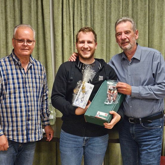 Drei Männer stehen vor einem grünen Vorhang. Der Mann in der Mitte hält eine grüne Box und ein verpacktes Geschenk und lächelt. Der Mann auf der linken Seite trägt ein kariertes Hemd, und der Mann auf der rechten Seite trägt ein blaues Hemd.
