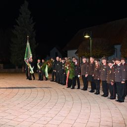Eine Gruppe von Menschen in Uniform steht nachts im Kreis und hält Blumen und eine Flagge. Hinter ihnen befindet sich ein Gebäude mit Dach.