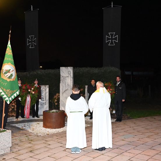 Eine nächtliche Gedenkzeremonie mit zwei Priestern, einer Flagge und Blumenarrangements vor einem Denkmal.