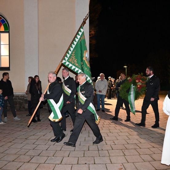 Männer in Uniform mit Fahnen gehen nachts durch einen gepflasterten Innenhof vor einem Gebäude. Mehrere Leute schauen zu.