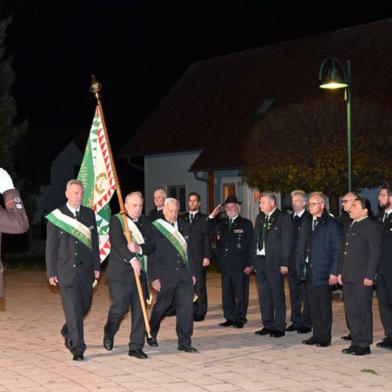 Bei Nacht ist eine Gruppe formell gekleideter Männer zu sehen, die vor einem Gebäude gehen. Sie tragen eine Fahne. Eine weitere Gruppe formell gekleideter Männer steht hinter ihnen, einige salutieren.