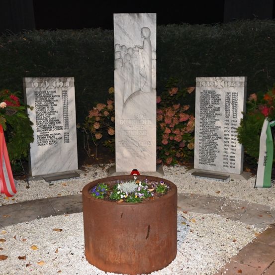 Drei Steinmonumente stehen in einem Garten. Sie listen Namen und Todesjahre auf. Ein zentraler Blumentopf mit Blumen ist davor platziert. Der Hintergrund zeigt Büsche und bunte Blumen.