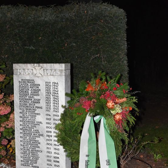 Ein Gedenkstein mit Namen und Daten von Personen, möglicherweise von einem Friedhof, ist mit einem Kranz verziert, der Blumen und Tannenzapfen enthält. Das Denkmal ist von Grün umgeben, und der Hintergrund ist dunkel.