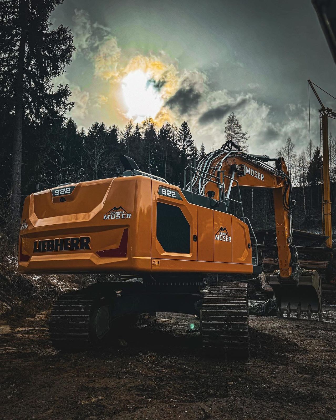 Ein orangefarbener Liebherr-Bagger mit der Nummer 922 steht in einem Waldgebiet, wobei die Sonne durch die Wolken scheint.