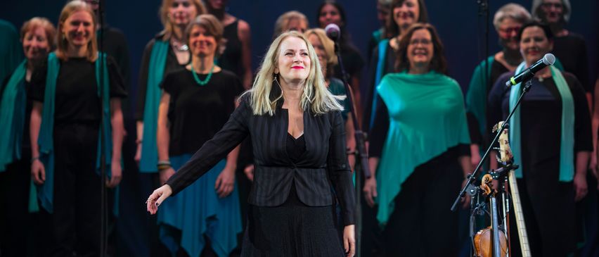 A woman stands on stage with a choir, both dressed in black. She smiles and gestures while the choir sings behind her. Microphones and a violin are present.