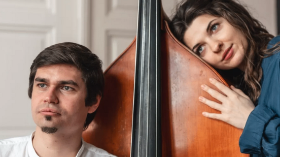 Ein Mann und eine Frau stehen neben einem großen Kontrabass, der Mann sieht ernst aus und die Frau lächelt mit der Hand auf dem Bass.