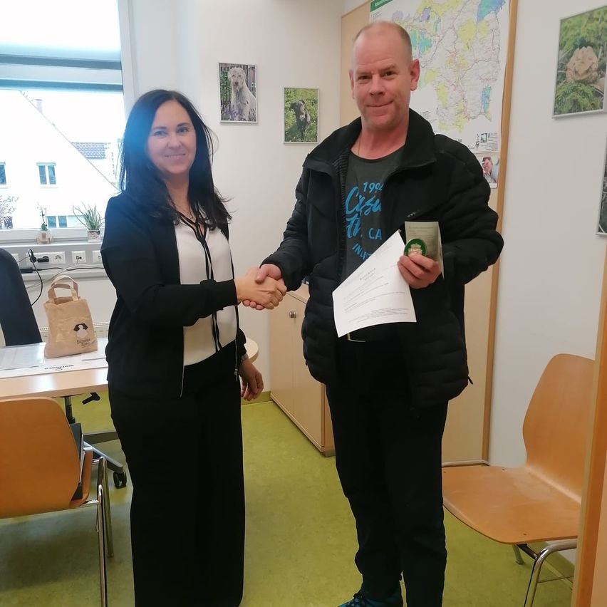 Ein Mann und eine Frau stehen in einem Raum mit Fenster und schütteln sich die Hände. Der Mann hält ein Zertifikat und die Frau lächelt.