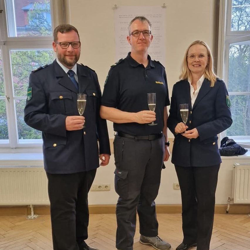 Drei uniformierte Personen, zwei Männer und eine Frau, stehen nebeneinander und halten Champagner-Gläser in der Hand. Der Mann auf der linken Seite trägt eine dunkelblaue Uniform, während der Mann in der Mitte und die Frau schwarze Uniformen tragen. Sie befinden sich in einem Raum mit einem Heizkörper an der Wand und einem Fenster mit Blick auf Bäume draußen.
