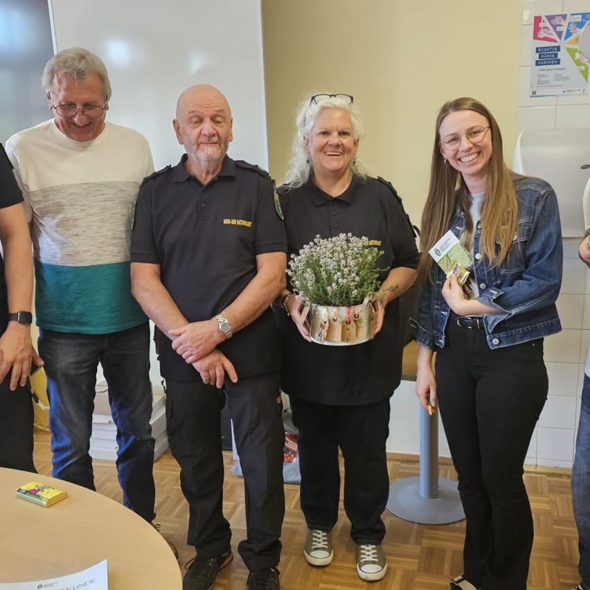 A group of individuals, including police officers and a woman holding flowers, stand together in a room. They are smiling and appear to be celebrating. A desk is in the foreground.