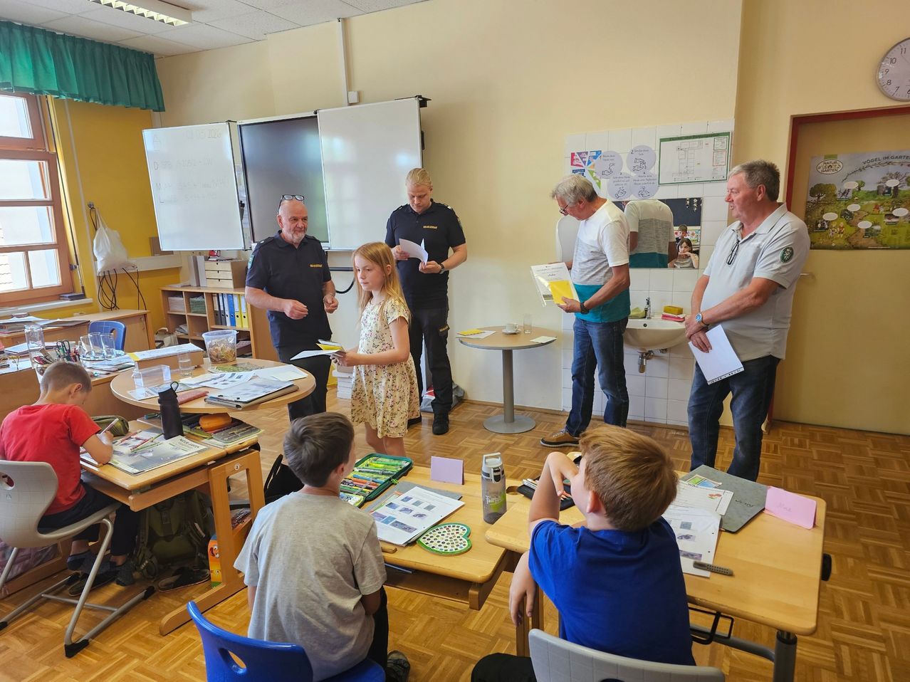 In a classroom, a young girl presents a piece of paper to a group of adults, while others read papers. Desks are filled with books and papers. A whiteboard and monitors are mounted on the wall.