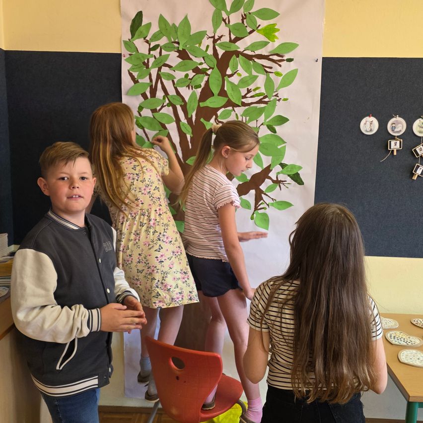 Four children are in a room, with three girls standing and one boy looking at a tree poster. The tree has green leaves and a brown trunk. The girls seem to be doing an art project.