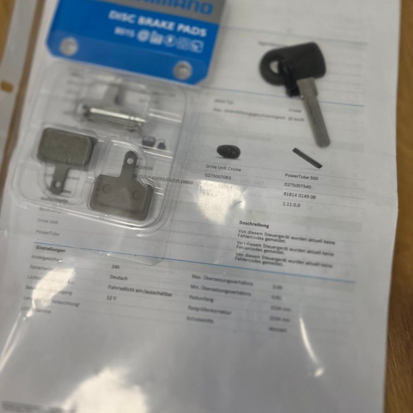 A package of Shimano disc brake pads is opened on a white paper with a key and a small metal piece. The package has a blue label with the text 'DISC BRAKE PADS'. The paper has a table with information about the product.