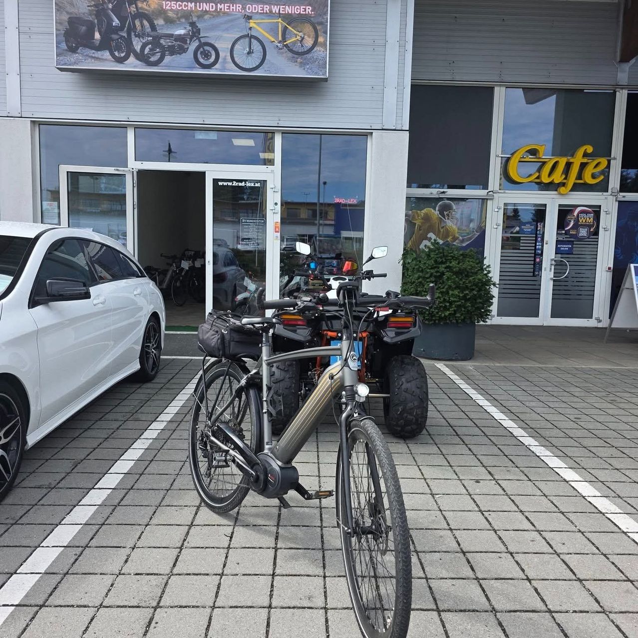 A white car and a bicycle are parked in front of a store with a sign that reads Cafe. The store has glass doors and windows, and there is a poster on the wall with a picture of a bicycle.