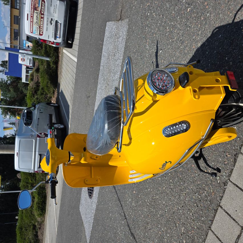 A bright yellow Vespa scooter is parked on the side of the road with a white van behind it.