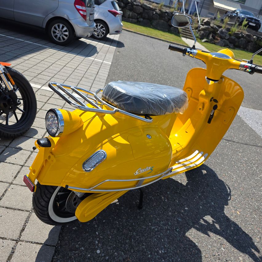 A bright yellow Vespa scooter with a gray seat cover is parked on a paved road with other vehicles in the background.