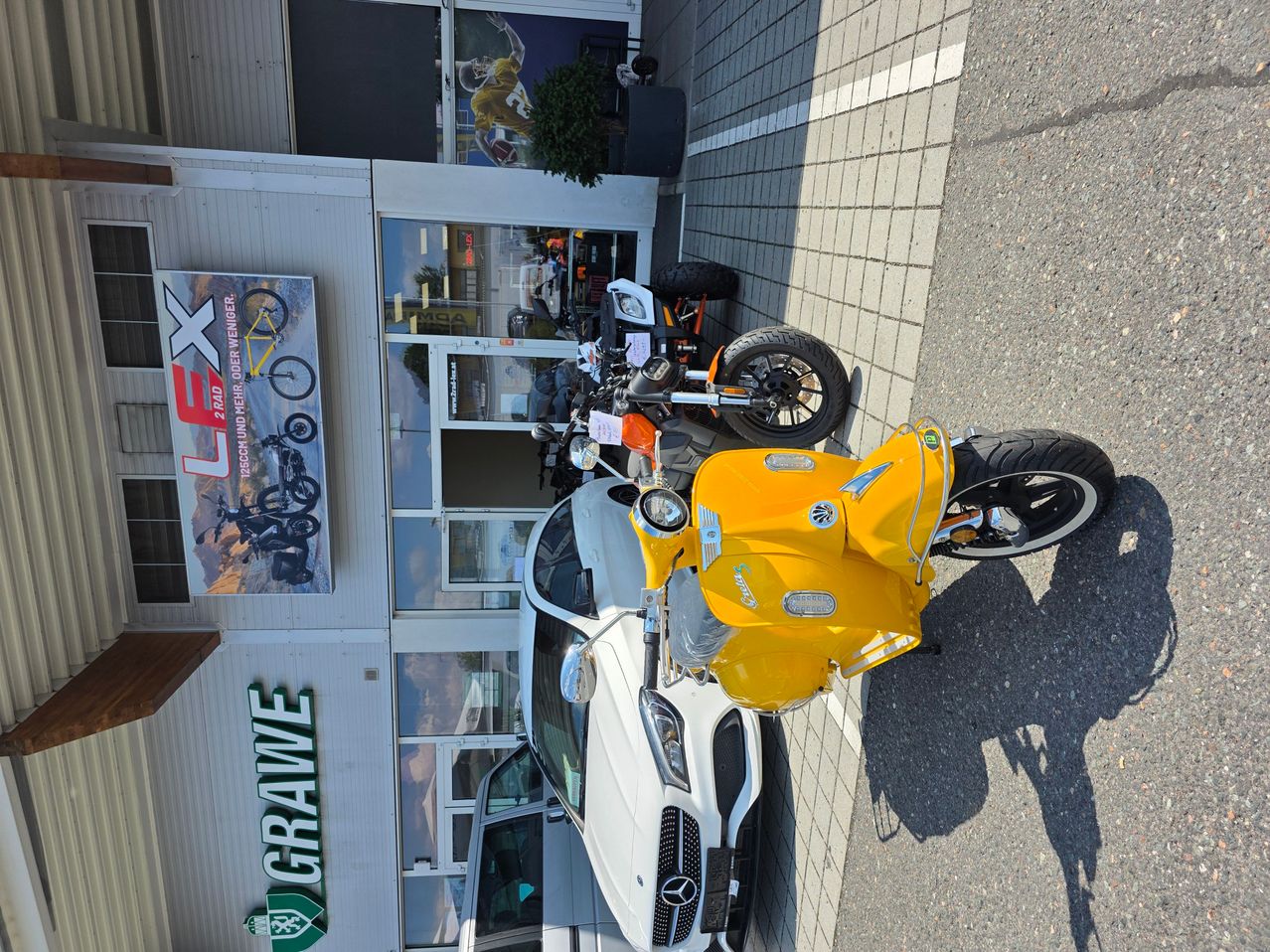 A yellow scooter is parked in front of a building with a sign that reads 'Grave' and 'LX'. A white car is parked next to it. The building has glass windows and a door. There are other motorcycles parked on the side.
