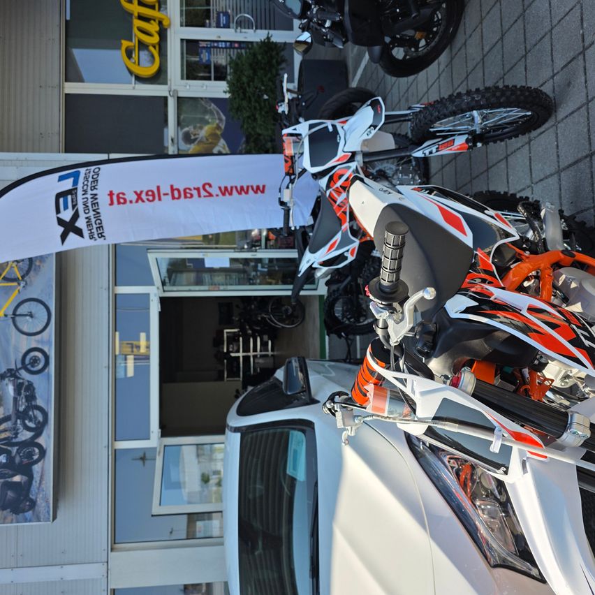 Ein weißer und orangefarbener Motorrad ist vor einem Gebäude mit einem Banner mit der Aufschrift 'TEX' und einer Website-Adresse geparkt. Ein weißes Auto ist daneben geparkt.